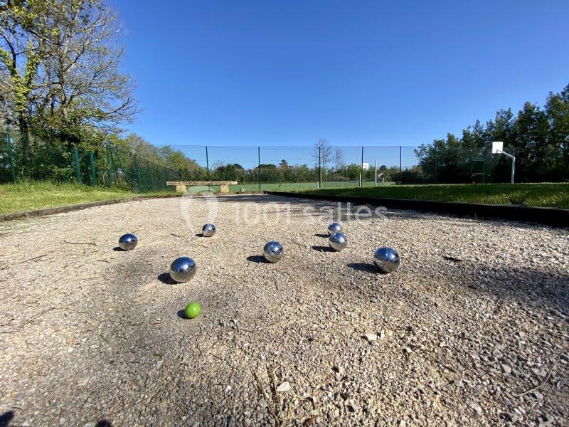 Terrain de pétanque avec plusieurs boules métalliques et un cochonnet vert, entouré de verdure et d'un terrain de sport.