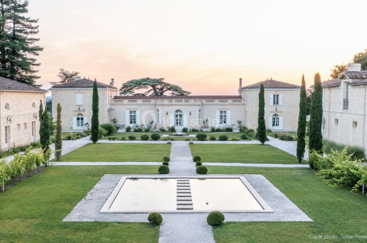 Vue d'un domaine avec jardin symétrique, bassin central, allées gravillonnées et bâtiments en pierre sous un ciel clair.
