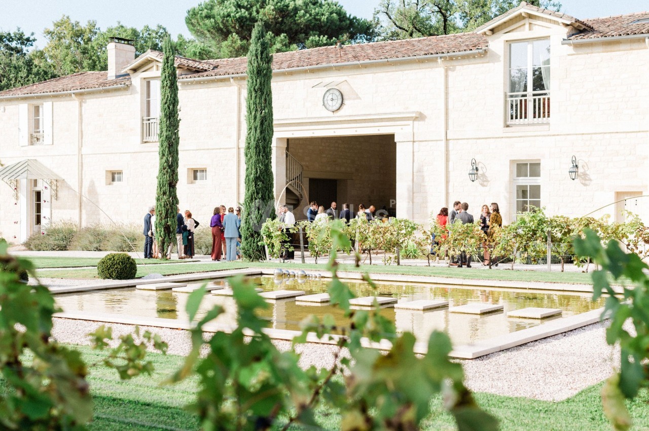 Des personnes rassemblées devant un bâtiment en pierre clair, entouré de jardins avec un bassin et des vignes au premier…