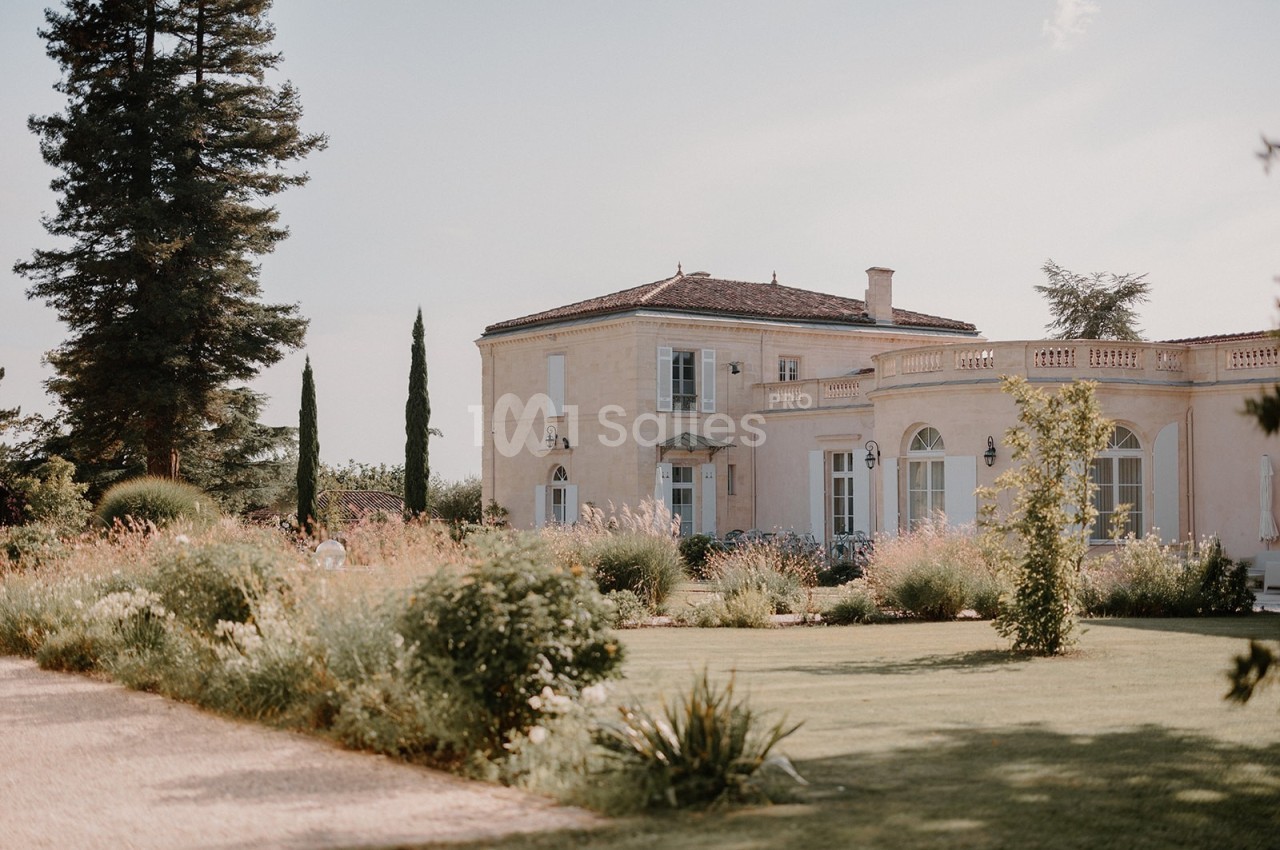 Grande maison en pierre entourée d'un jardin fleuri et arboré, sous un ciel clair.