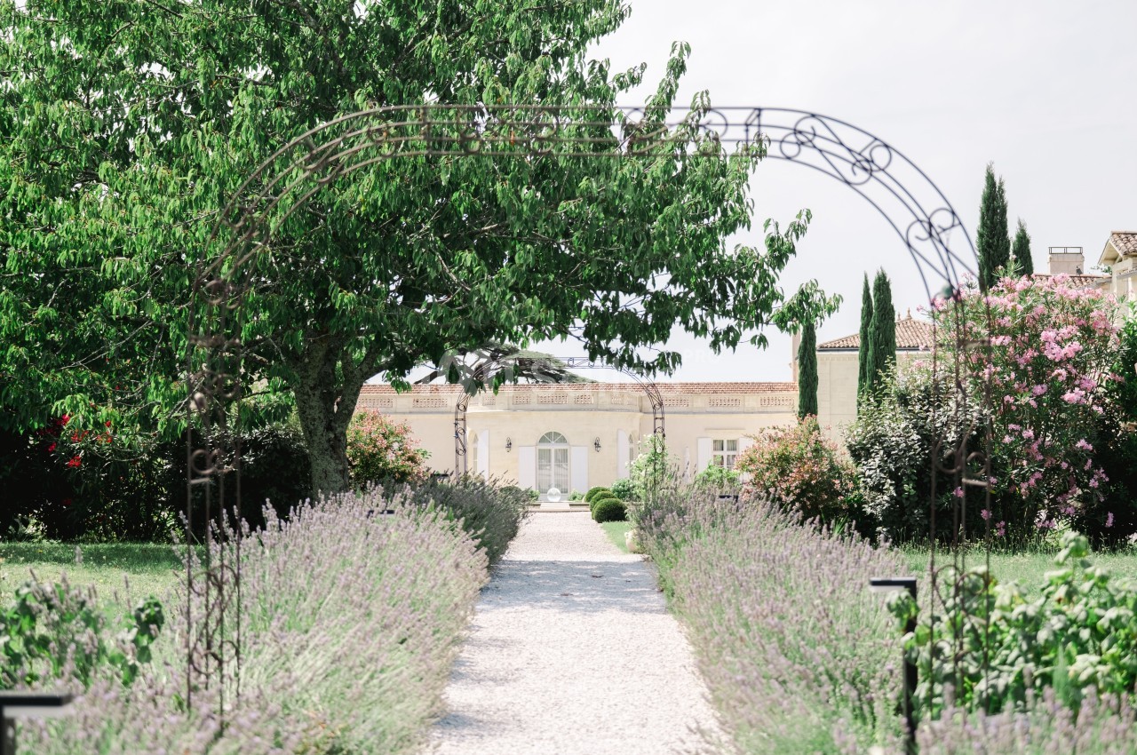 Allée bordée de lavande menant à une maison en pierre claire, encadrée par des arbres et des arches métalliques.