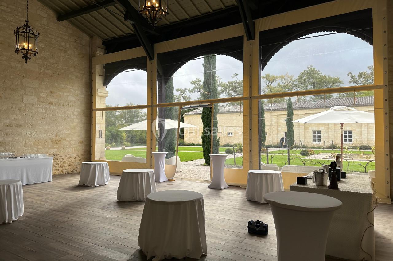 Salle lumineuse avec tables hautes drapées de blanc, grandes baies vitrées donnant sur un jardin avec parasols.