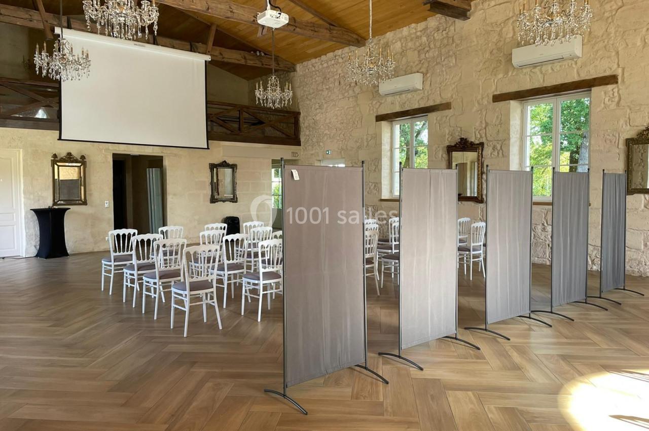 Salle lumineuse avec parquet, chaises blanches disposées en rangées, écrans de séparation et projecteur au plafond.