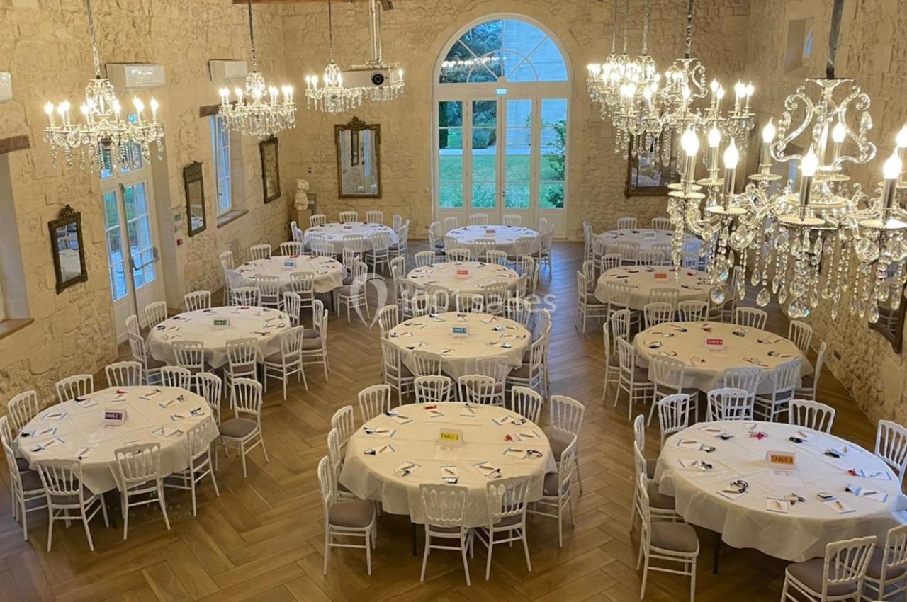Salle de réception avec tables rondes dressées, chaises blanches, lustres en cristal et grandes fenêtres donnant sur un…