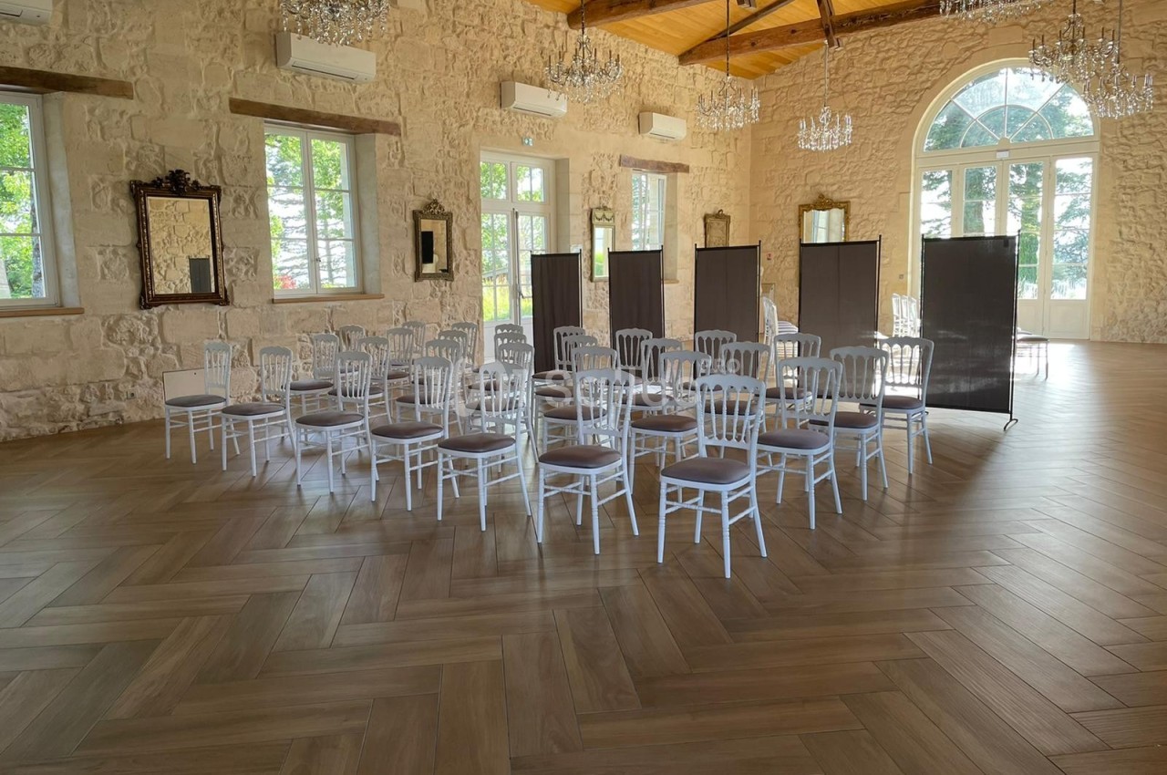 Salle lumineuse avec parquet en bois, chaises blanches disposées en cercle et paravents noirs devant des fenêtres.