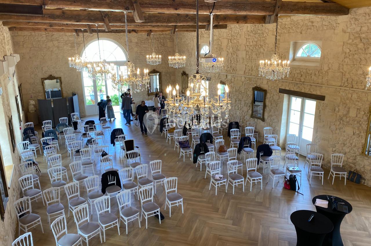 Salle en pierre avec des chaises blanches disposées pour un événement, éclairée par des lustres et de grandes fenêtres.