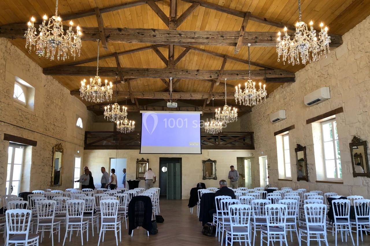 Salle de réception avec poutres apparentes, lustres, chaises blanches alignées et écran de projection allumé.