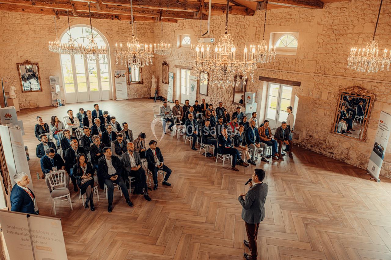 Un conférencier s'adresse à un public assis dans une salle lumineuse avec des murs en pierre et des lustres.
