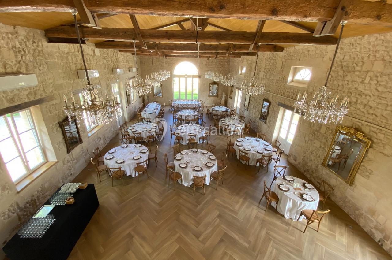 Salle de réception avec tables rondes dressées, parquet en bois, murs en pierre et lustres suspendus.