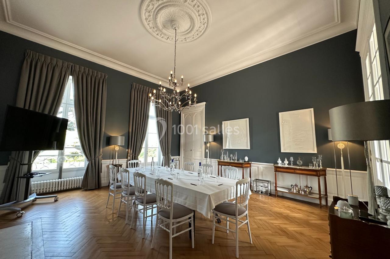 Salle à manger élégante avec table dressée, chaises blanches, lustre central, murs bleu foncé et parquet en bois clair.