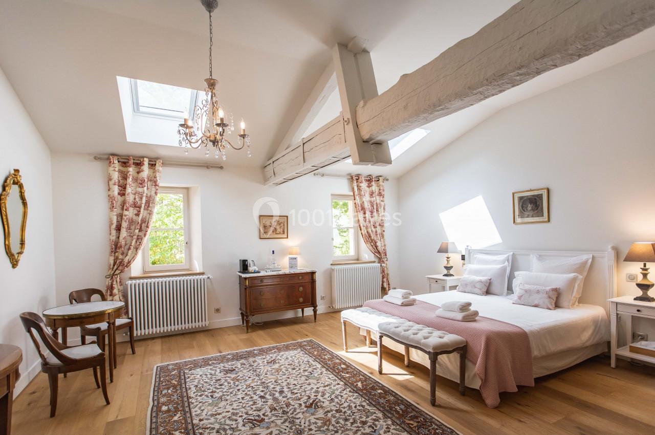 Chambre lumineuse avec lit double, poutres apparentes, tapis, mobilier classique et fenêtres laissant entrer la lumière…
