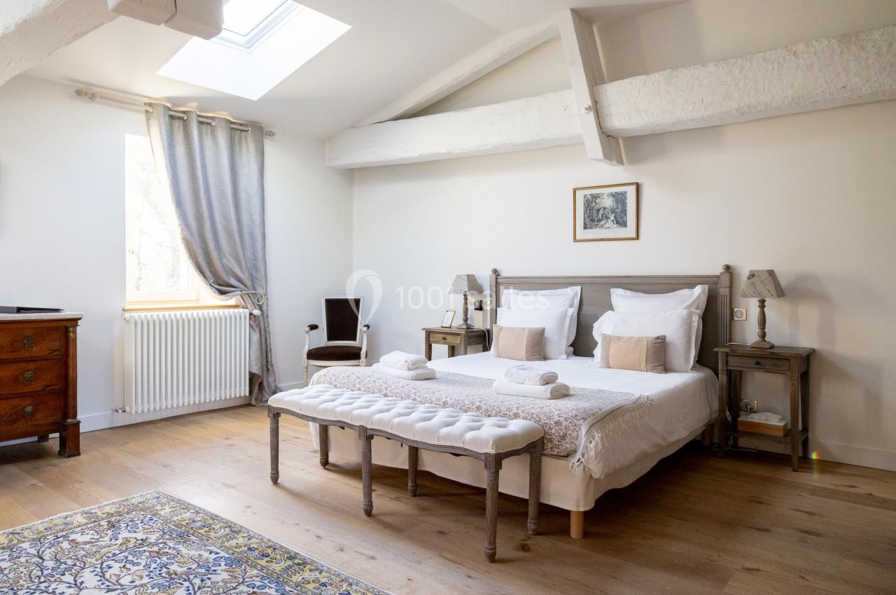 Chambre lumineuse avec lit double, poutres apparentes, parquet, tapis et fenêtre avec rideaux gris.