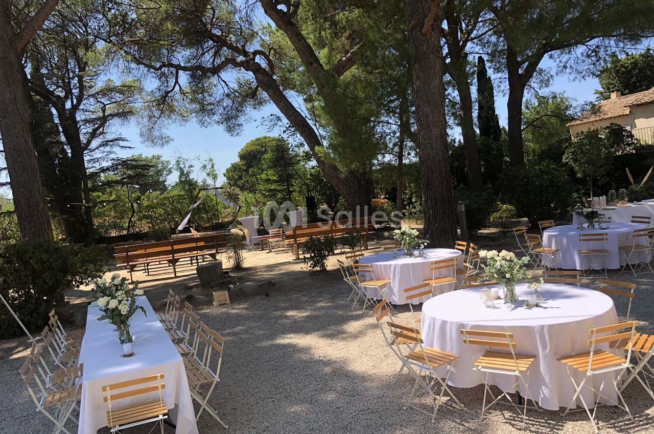 Tables rondes décorées de fleurs blanches et chaises en bois disposées en extérieur sous des arbres dans un cadre ensoleillé.