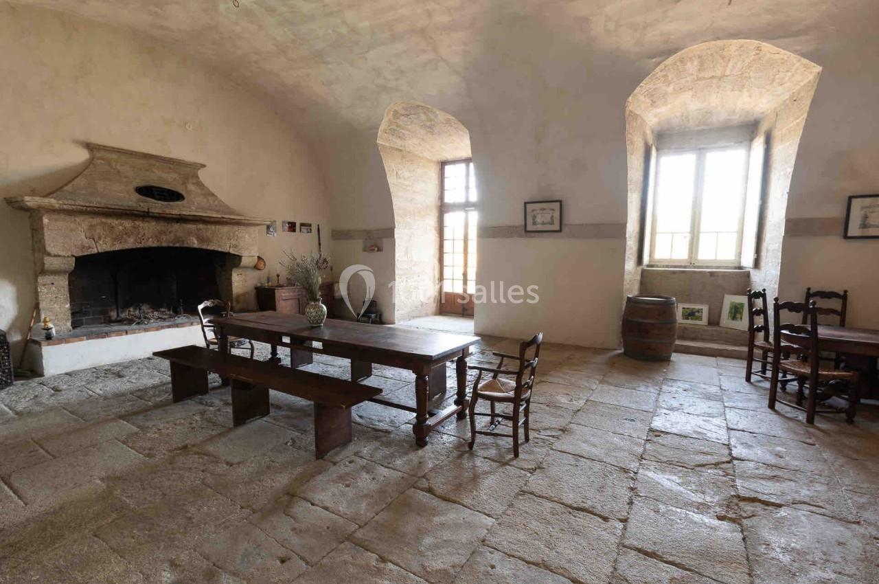 Salle rustique avec cheminée en pierre, grande table en bois, bancs et chaises, éclairée par des fenêtres à double battant.