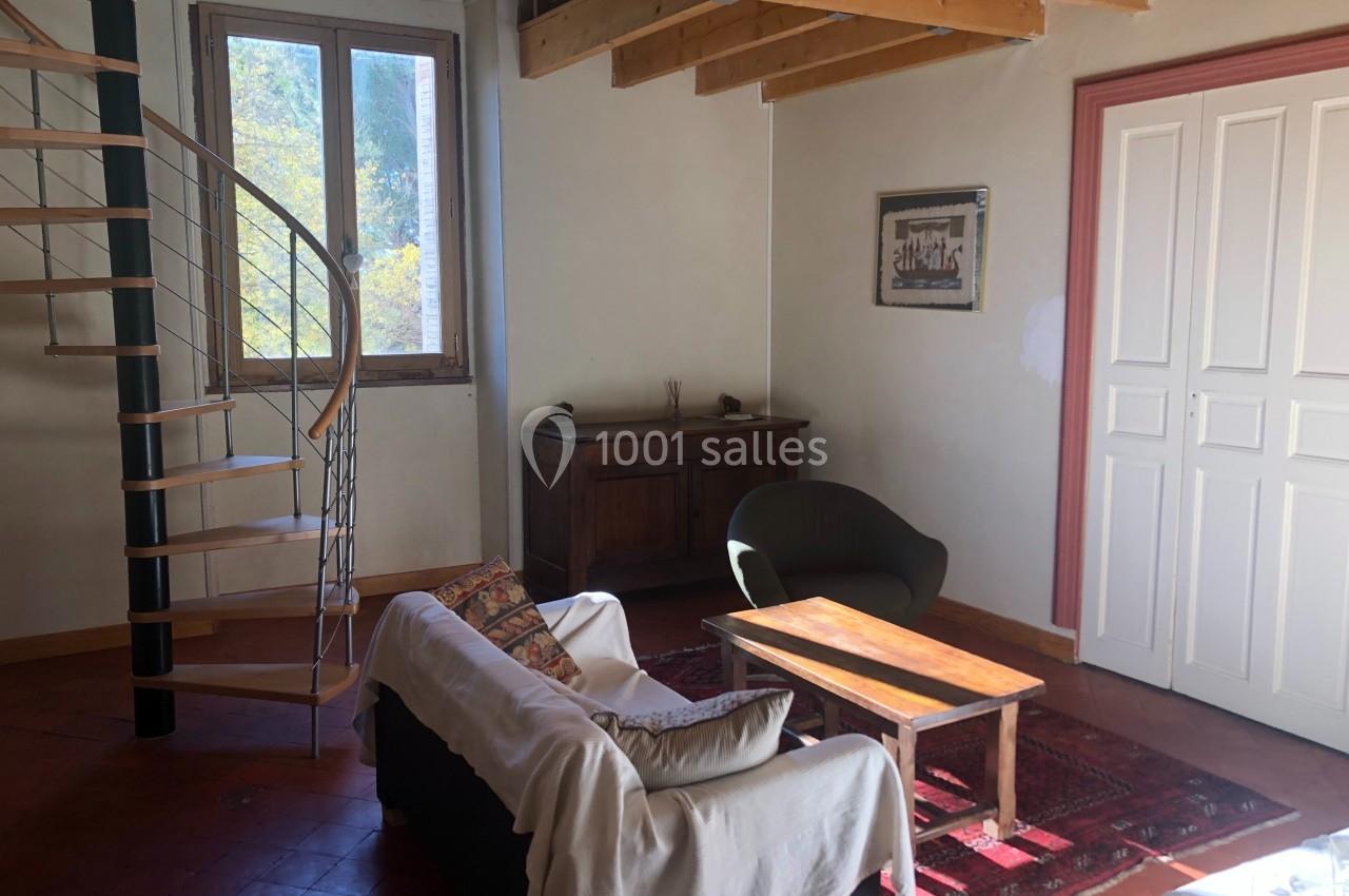 Salon lumineux avec canapé, fauteuil, table basse, escalier en colimaçon, poutres apparentes et fenêtre donnant sur un arbre.