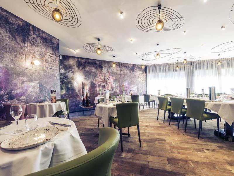 Salle de restaurant lumineuse avec tables dressées, chaises vertes, parquet en bois et décoration murale florale.