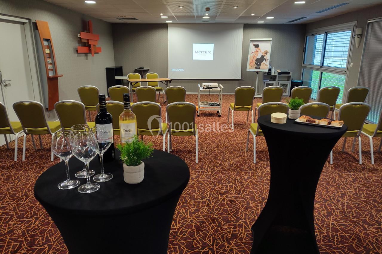 Salle de réunion équipée avec chaises, projecteur, tables hautes décorées, verres et bouteilles de vin.