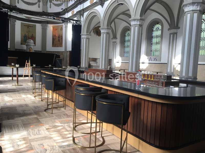 Intérieur d'une ancienne église transformée en bar, avec un comptoir moderne et des tabourets noirs.
