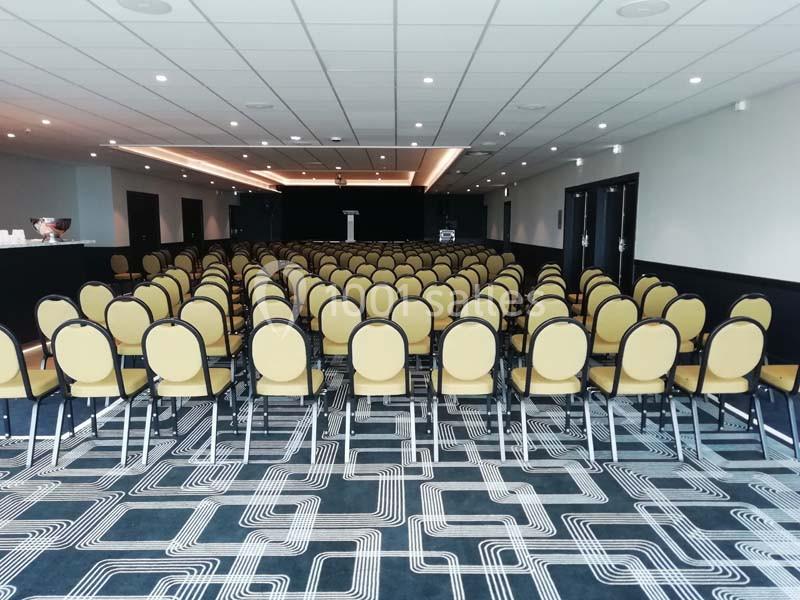 Salle de conférence avec rangées de chaises jaunes alignées face à une estrade vide.