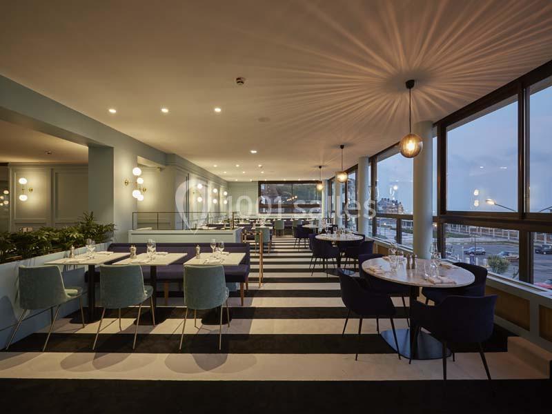 Salle de restaurant moderne avec tables dressées, chaises colorées et grandes baies vitrées donnant sur l'extérieur.
