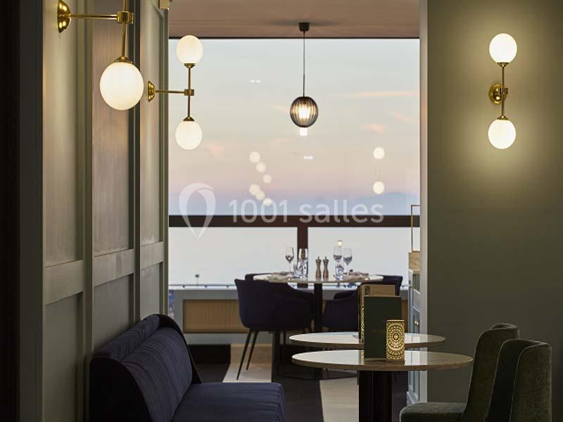 Salle de restaurant élégante avec vue sur un coucher de soleil, éclairée par des appliques murales et des suspensions.