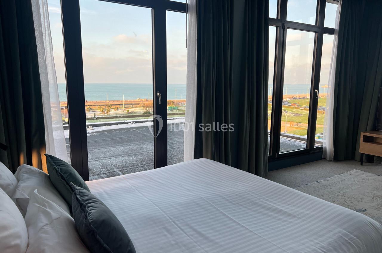 Vue depuis une chambre avec un lit, grandes baies vitrées et rideaux ouverts donnant sur un paysage côtier.