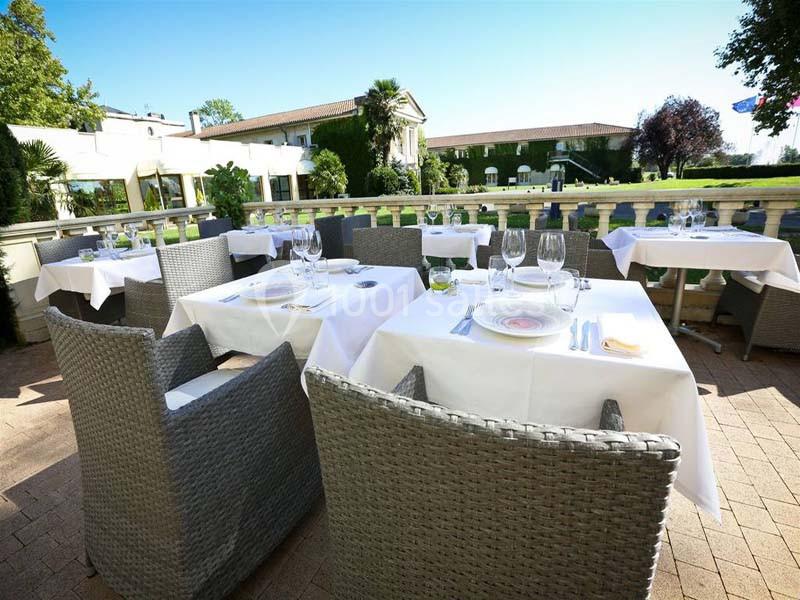 Terrasse d'un restaurant avec tables dressées, nappes blanches et chaises en rotin, entourée de verdure.