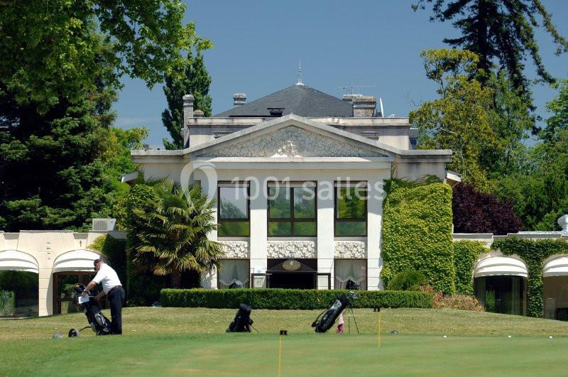 Façade d'un bâtiment entouré de végétation, avec un golfeur et des sacs de golf au premier plan.