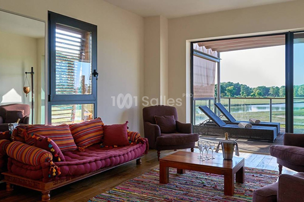 Salon lumineux avec canapé coloré, fauteuils, table basse en bois, grande baie vitrée et vue sur un lac verdoyant.