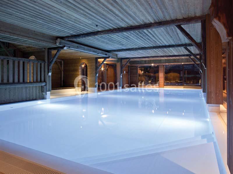 Piscine intérieure éclairée entourée de structures en bois sous un plafond à poutres apparentes.