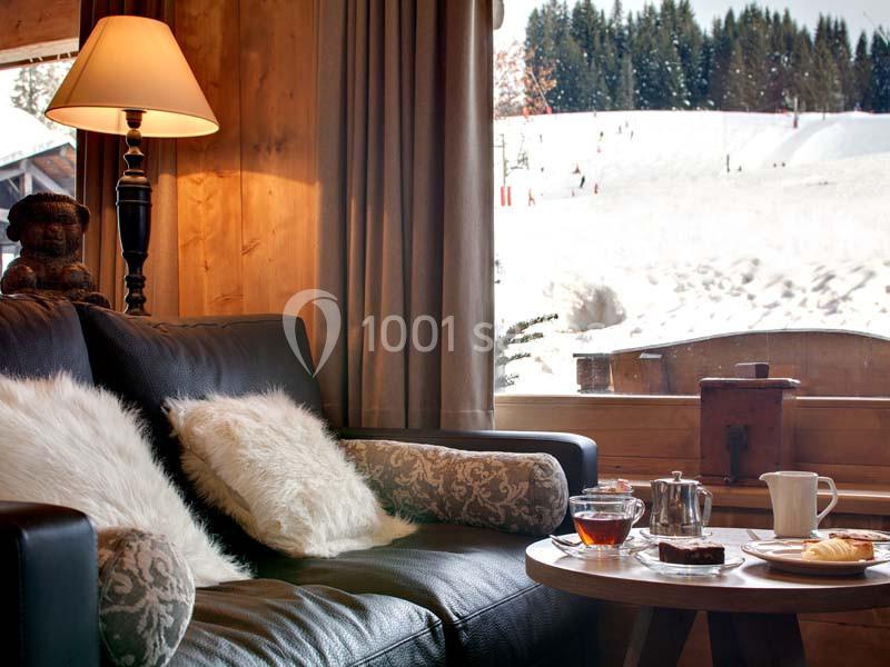Salon chaleureux avec canapé, coussins, table basse et vue sur un paysage enneigé à travers une grande fenêtre.