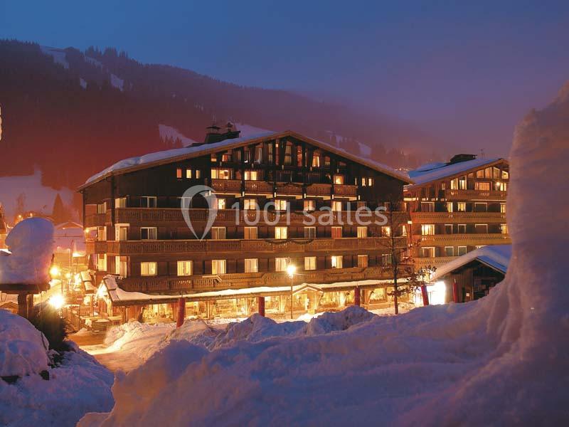 Chalet éclairé en soirée, entouré de neige, avec des montagnes en arrière-plan sous un ciel crépusculaire.