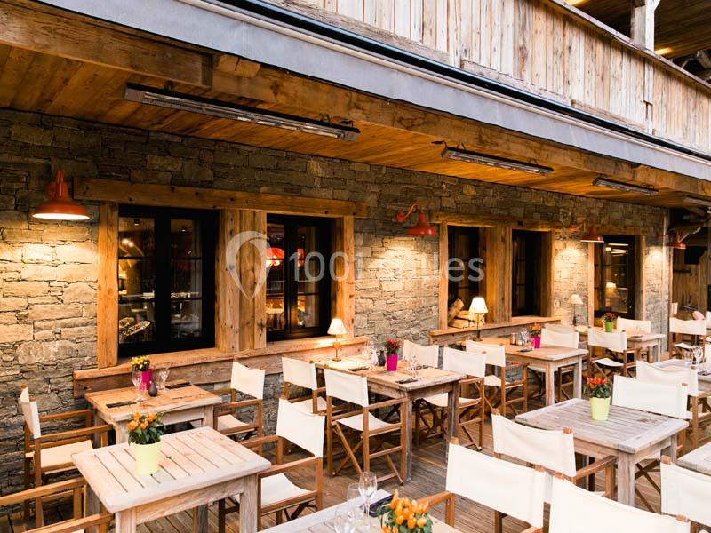 Terrasse en bois avec tables et chaises blanches, décorée de plantes en pots, devant une façade en pierre éclairée.