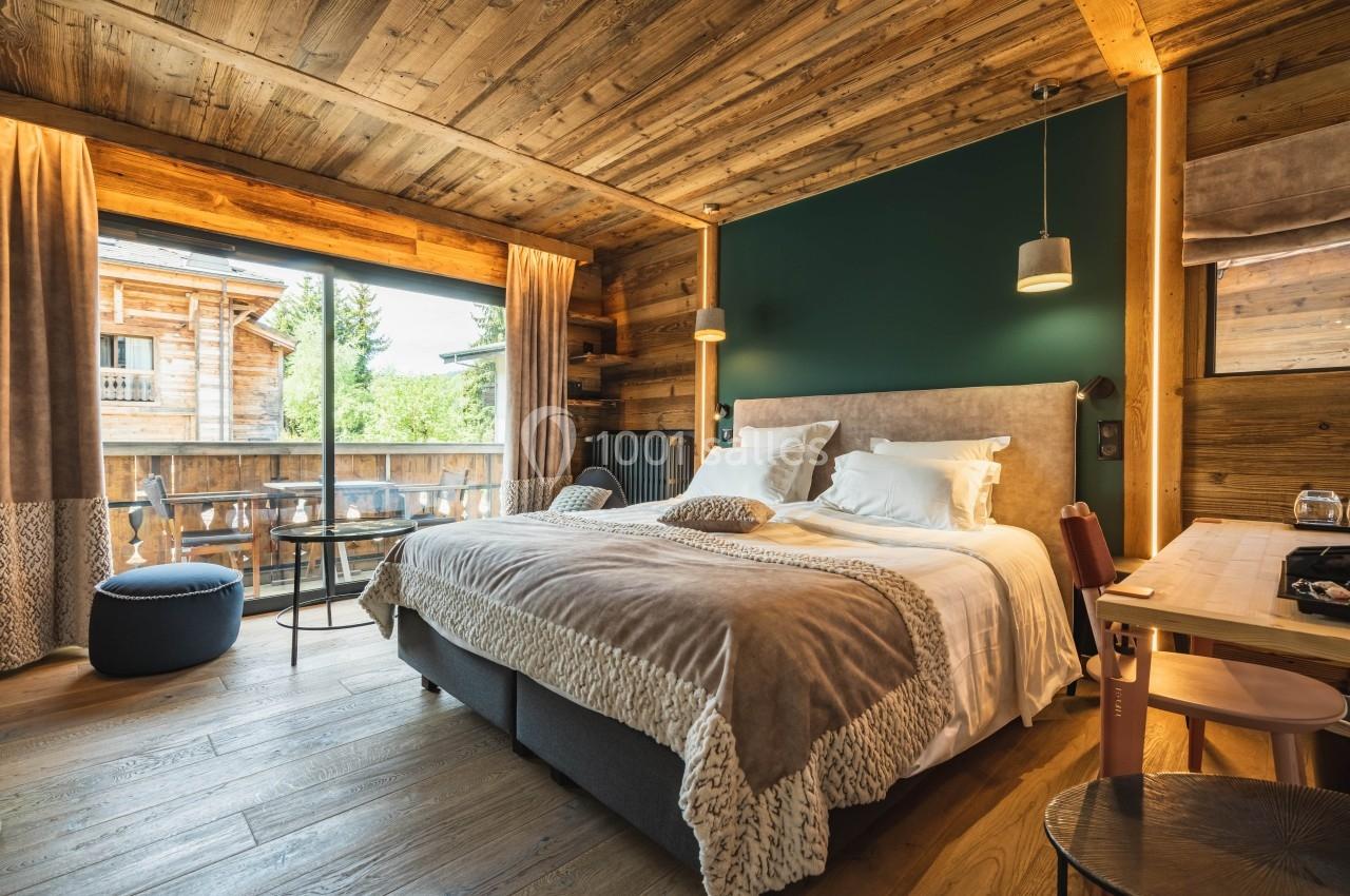 Chambre lumineuse avec lit double, décor en bois, grande baie vitrée donnant sur un balcon et vue sur la nature.