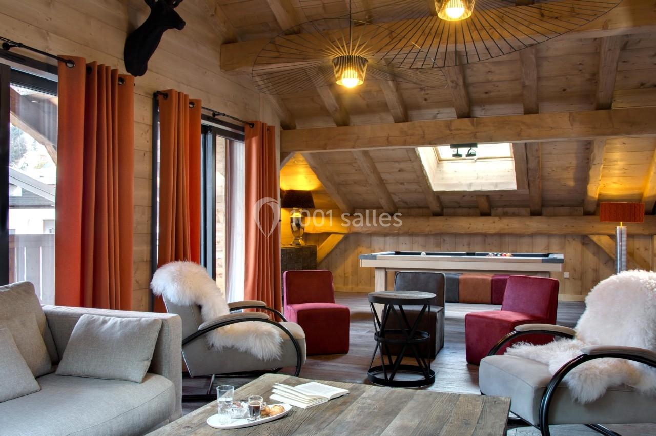 Salon chaleureux sous combles avec poutres apparentes, canapé, fauteuils, table basse en bois et table de billard.