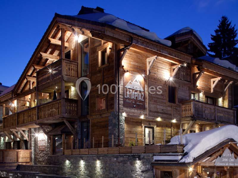 Chalet en bois éclairé de nuit, avec balcons et toit enneigé, entouré d'arbres dans un cadre montagnard.