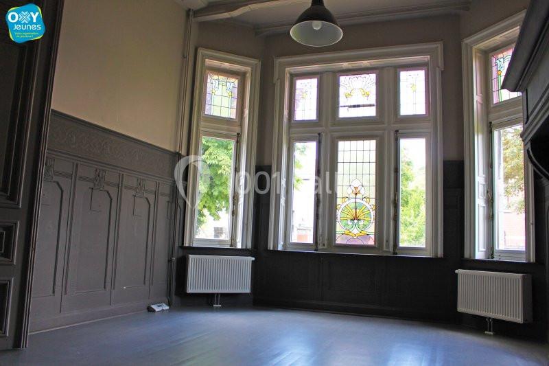 Location salle Farciennes (Hainaut) - Château Oxyjeunes #10 Salle vide avec parquet sombre, murs ornés de moulures et grandes fenêtres avec vitraux colorés laissant entrer la lumière.