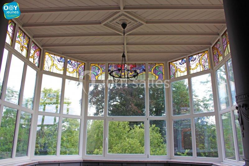 Location salle Farciennes (Hainaut) - Château Oxyjeunes #6 Baie vitrée avec vitraux colorés en partie haute, offrant une vue sur des arbres et un ciel dégagé.