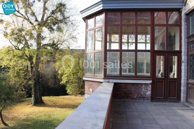 Location salle Farciennes (Hainaut) - Château Oxyjeunes #12 Véranda vitrée donnant sur un jardin arboré avec pelouse, vue depuis une terrasse en pierre.