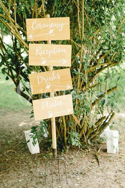 Panneau en bois avec flèches indiquant ’Ceremony’, ’Reception’, ’Drinks’ et ’Photos’, placé devant des arbustes.