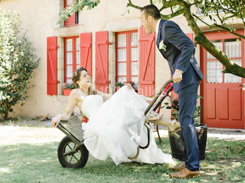Une mariée assise dans une brouette, poussée par un homme en costume, dans un jardin devant une maison aux volets rouges.