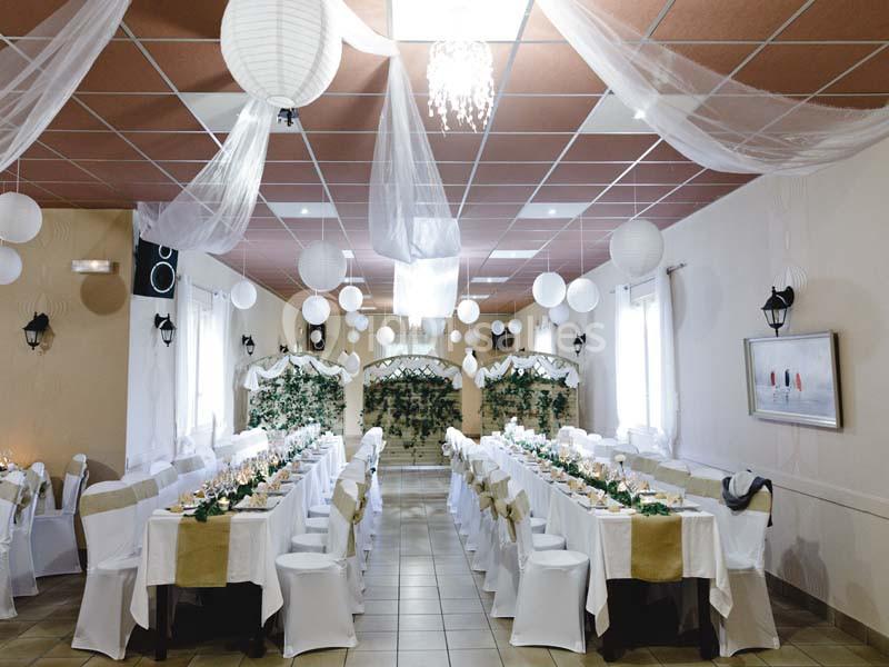Salle de réception décorée avec des tables alignées, nappes blanches, lanternes suspendues et guirlandes végétales.