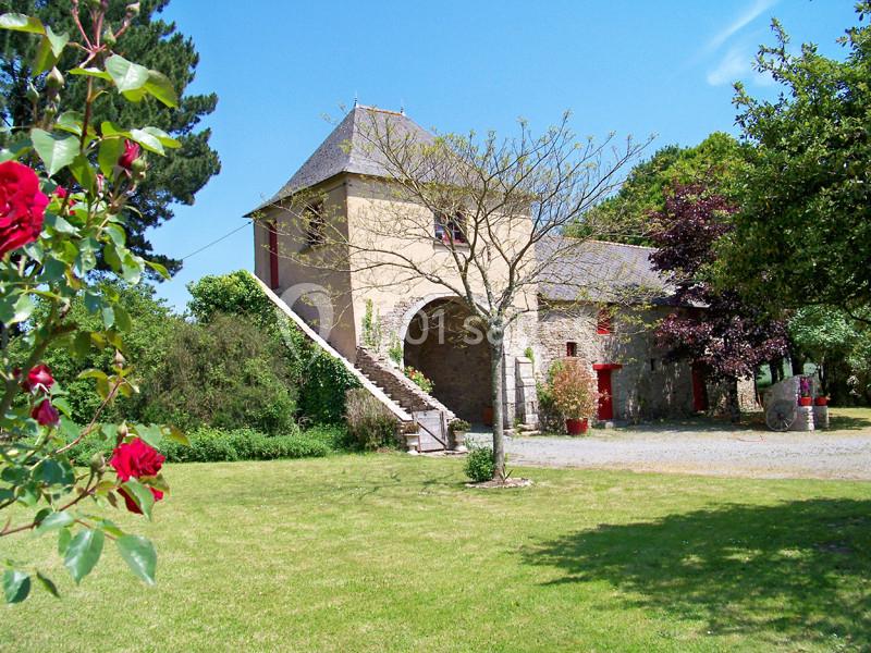 Bâtisse en pierre avec escalier extérieur, entourée de verdure et de fleurs sous un ciel bleu.