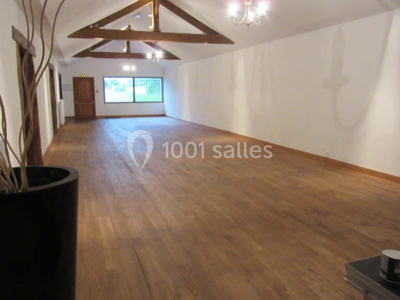 Salle vide avec parquet en bois, poutres apparentes au plafond, murs blancs et grande fenêtre au fond.