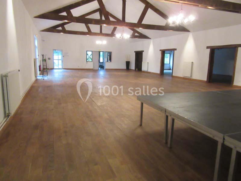 Salle spacieuse avec parquet en bois, poutres apparentes au plafond, éclairage suspendu et une petite estrade.