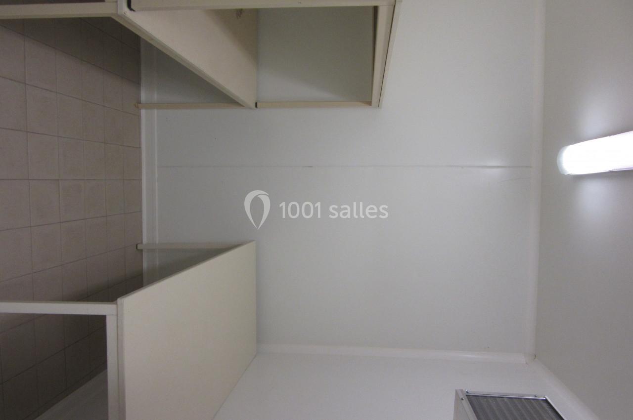 Cabine vide avec cloisons blanches, sol carrelé beige et éclairage fluorescent au mur.