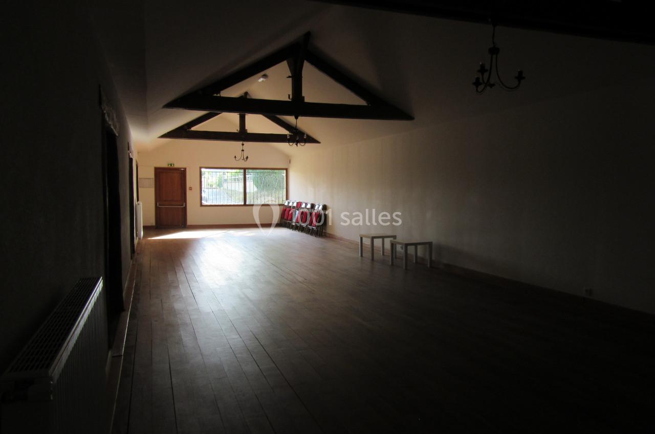 Salle vide avec parquet en bois, poutres apparentes au plafond, chaises empilées et lumière naturelle entrant par une…