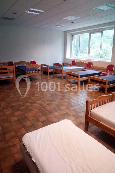 Salle avec plusieurs lits simples en bois, matelas bleus et rouges, sol en carrelage et grande fenêtre donnant sur des…
