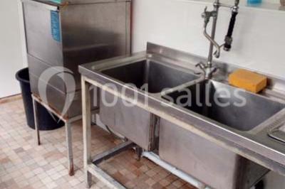 Miniature Location salle Trégomeur (Côtes-d'Armor) - Les Serres du Rodo #16 Évier double en inox avec robinet flexible, lave-vaisselle professionnel et sol en carrelage beige dans une cuisine.