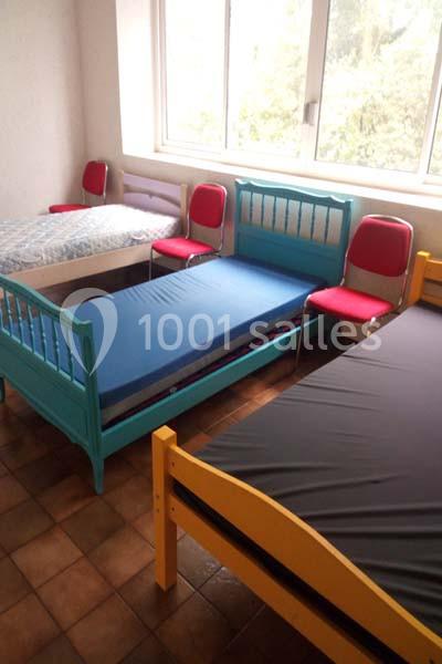Trois lits simples colorés et des chaises rouges disposés dans une pièce lumineuse avec un sol en carrelage.